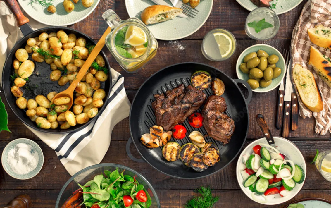 Stół pełen przygotowanych potraw ze świeżych warzyw, patelnia z grillowanym mięsem, pomidorami, cebulą, czosnkiem, cukinią i inne potrawy.